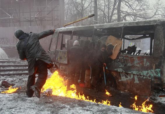 Pro-European protesters take cover behind a burnt bus as a demonstrator throws a stick during clashes with riot police in Kiev